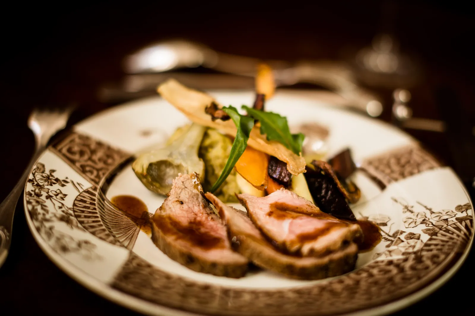 Gourmet plate from La Table de Siaurac in Bordeaux featuring sliced roasted meat with grilled vegetables and edible garnish, presented on classically decorated porcelain.