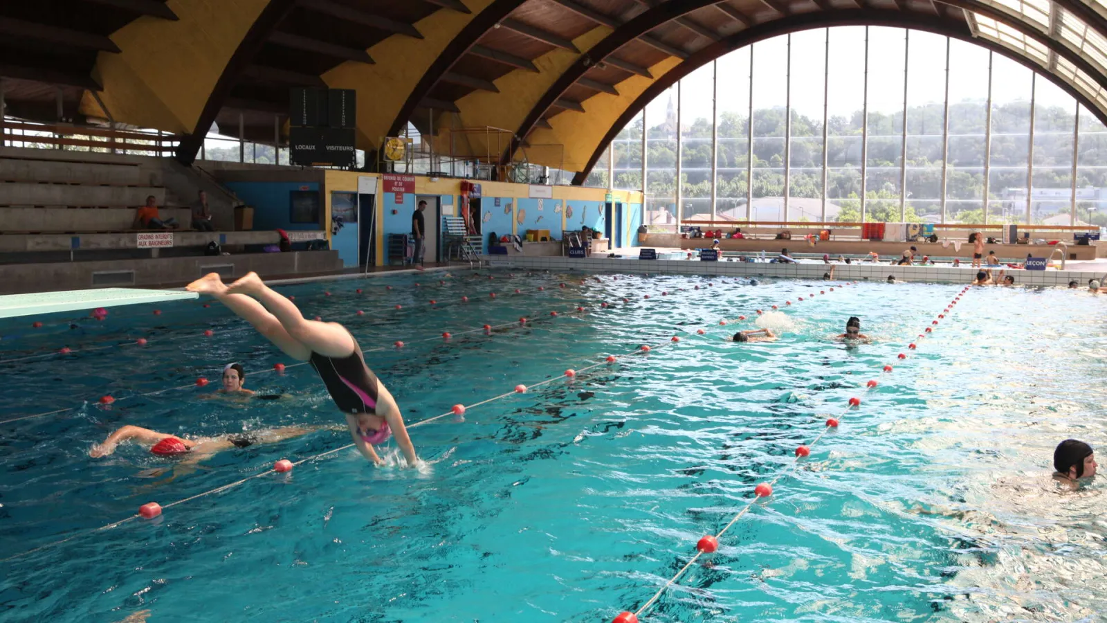 Interior view of Bordeaux's municipal swimming pool Galin with multiple swimmers and a diver in mid-dive, under a yellow vaulted roof and large windows overlooking the city