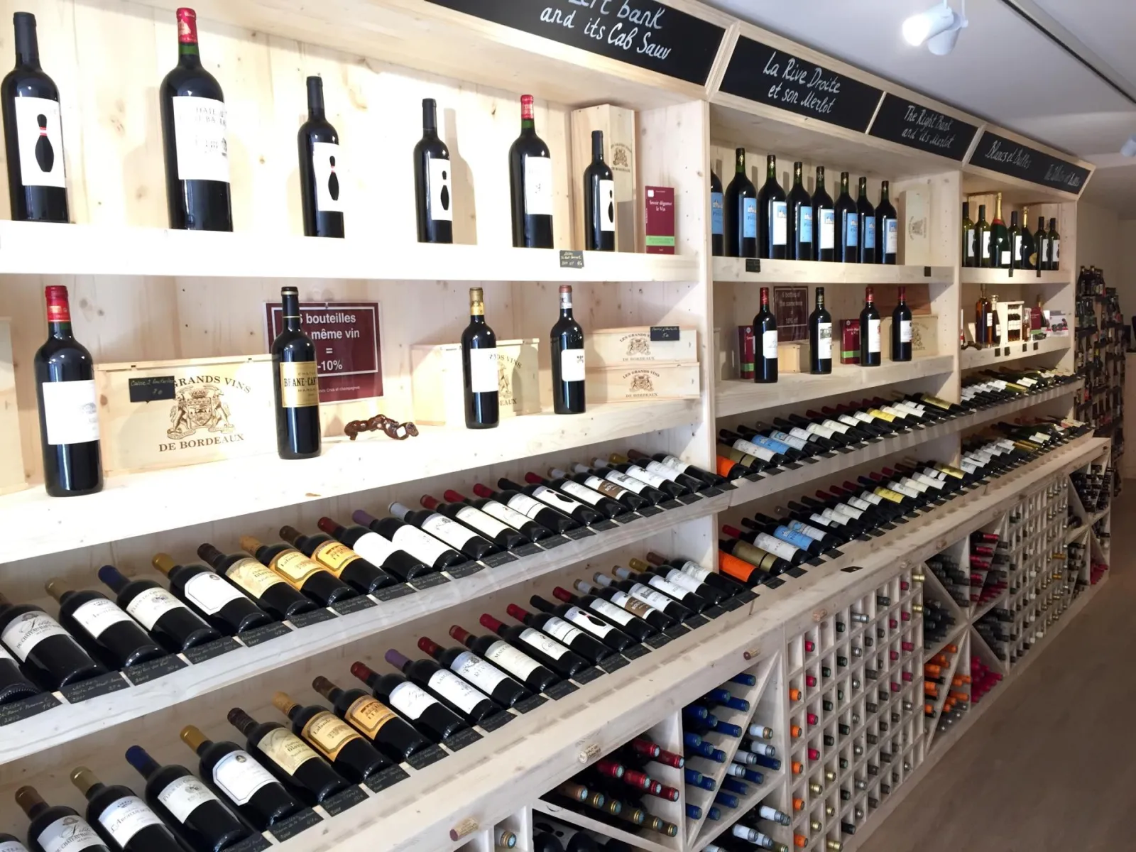 Wine shop display with Bordeaux bottles arranged on white shelves, featuring red wines, various vintages and promotional offers in a Bordeaux wine store.