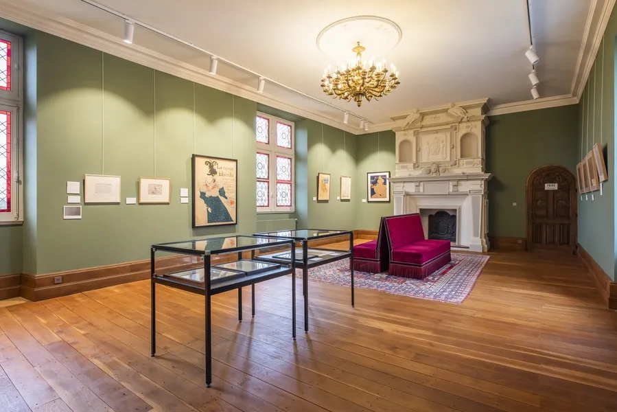 Exhibition gallery in Toulouse-Lautrec Castle in Bordeaux featuring sage green walls, wooden flooring, display cases, framed artworks, white fireplace, golden chandelier and burgundy furniture