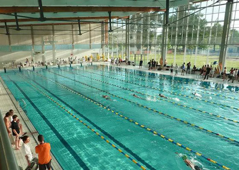 Interior view of the Olympic swimming pool of Villenave d'Ornon in Bordeaux with multiple swimmers in the lanes, spectators in the stands, and a large covered facility with metal structure.