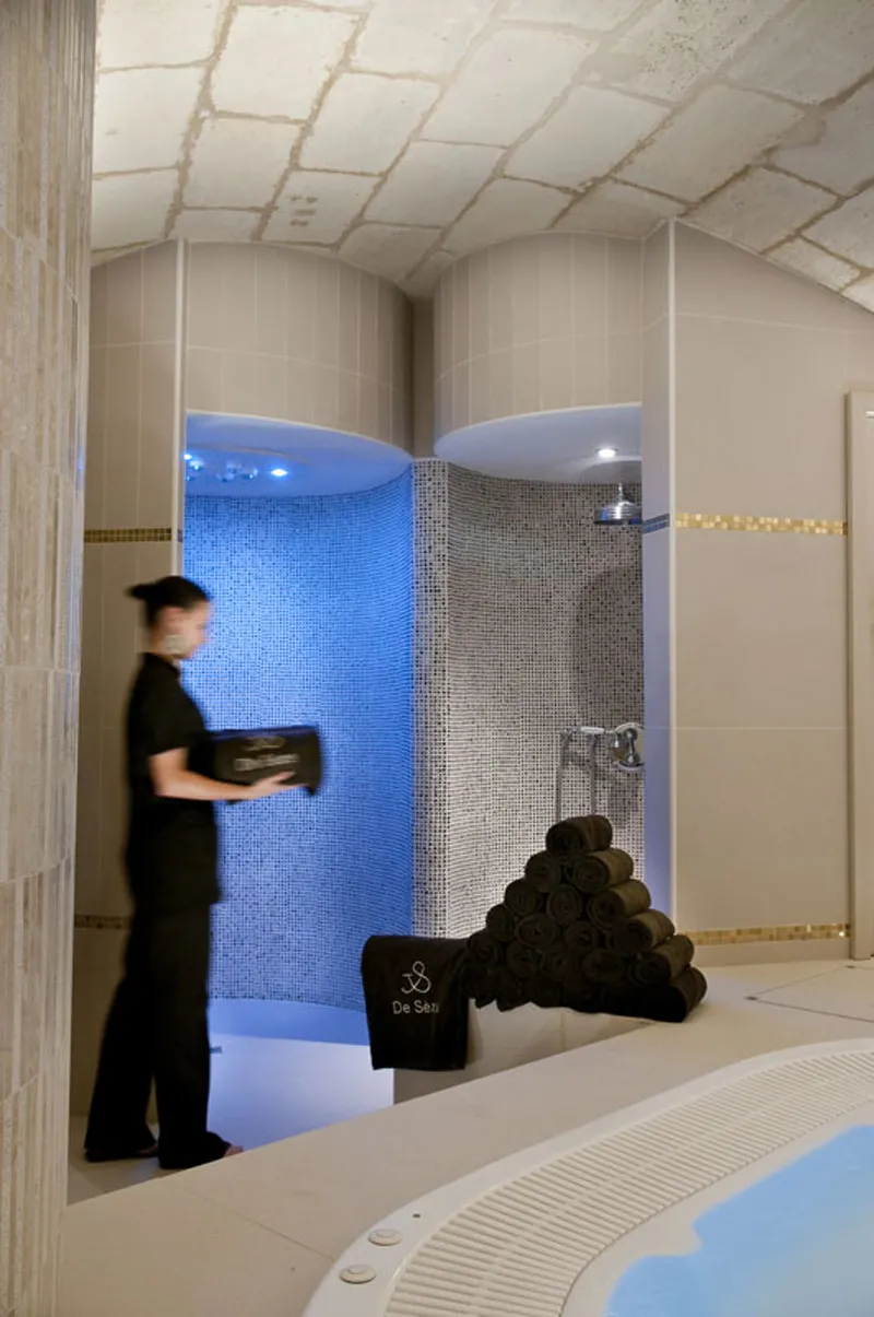 Interior of Hotel de Sèze spa in Bordeaux featuring a modern shower area with blue and gray tiled walls under ambient lighting, staff member in black uniform, and stacked black towels