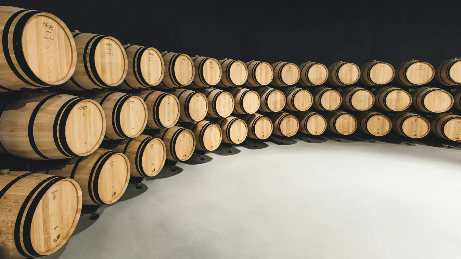 Rows of wooden barrels with black metal bands arranged in a winery aging cellar, lit by natural light against a minimalist gray and black background.