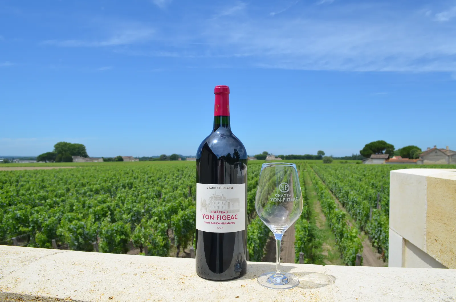 Château Yon-Figeac Grand Cru Classé bottle with wine glass overlooking the lush Saint-Émilion vineyard landscape in Bordeaux.