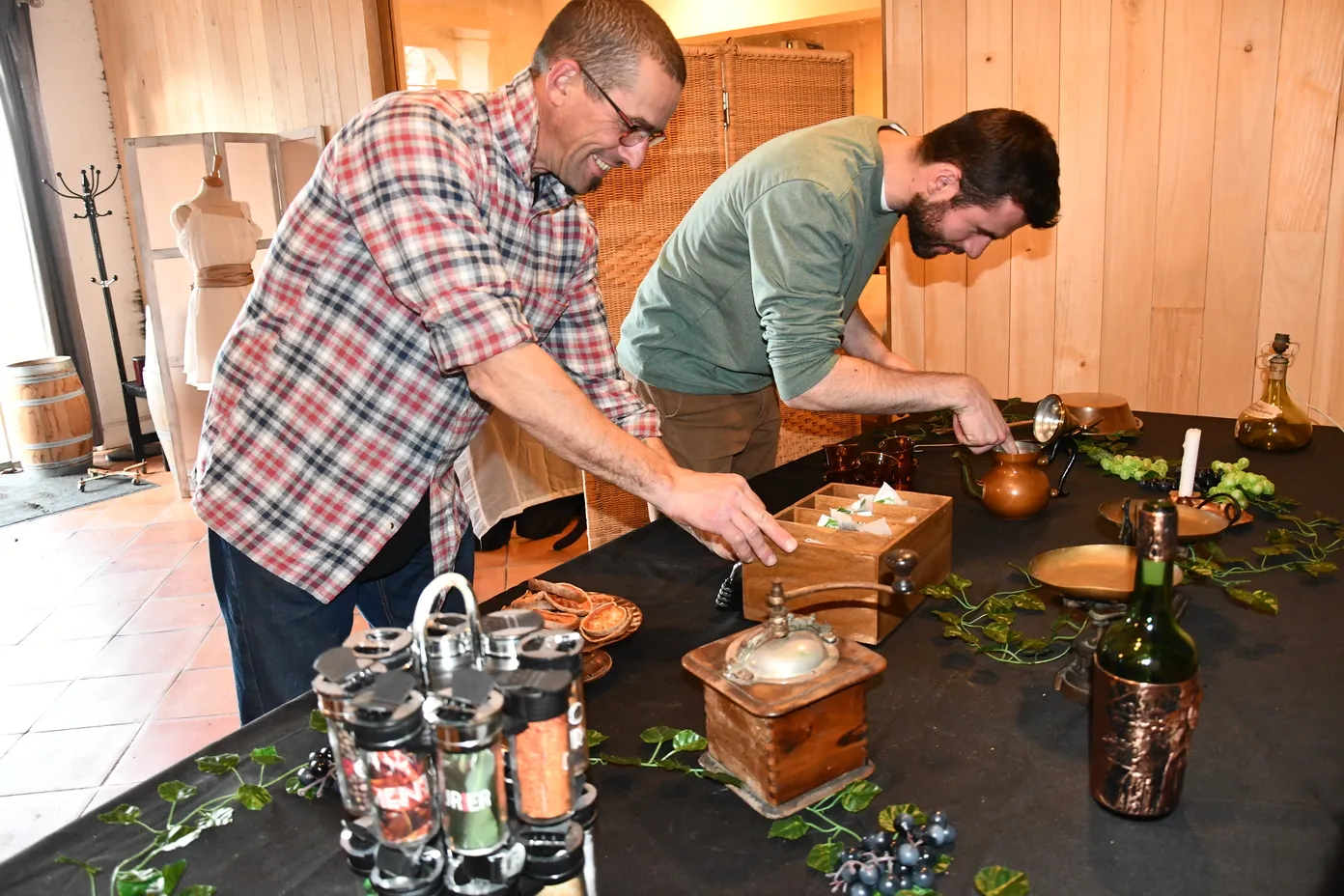 Two men participate in a wine tasting session in a wooden cellar. One pours wine into a glass while the other observes, surrounded by bottles, glasses and tasting accessories.