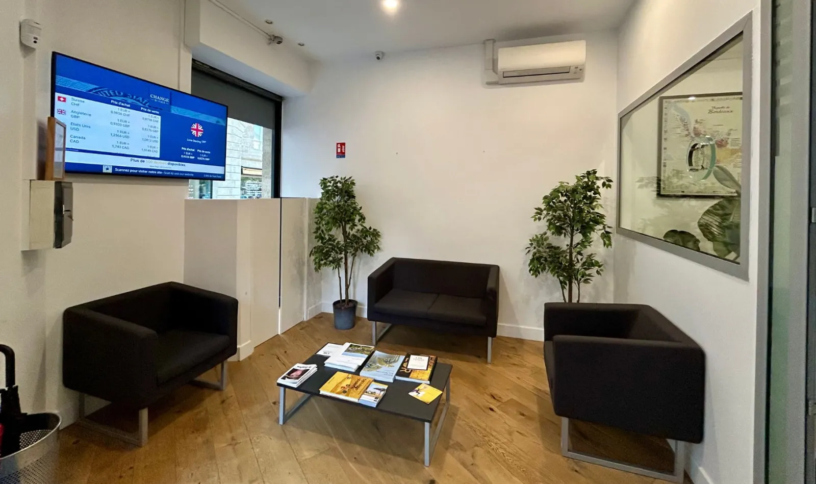 Modern waiting area of Fidso currency exchange office in Bordeaux, featuring three black couches, a coffee table with brochures, green plants, a currency rate display screen, and a glass reception desk.