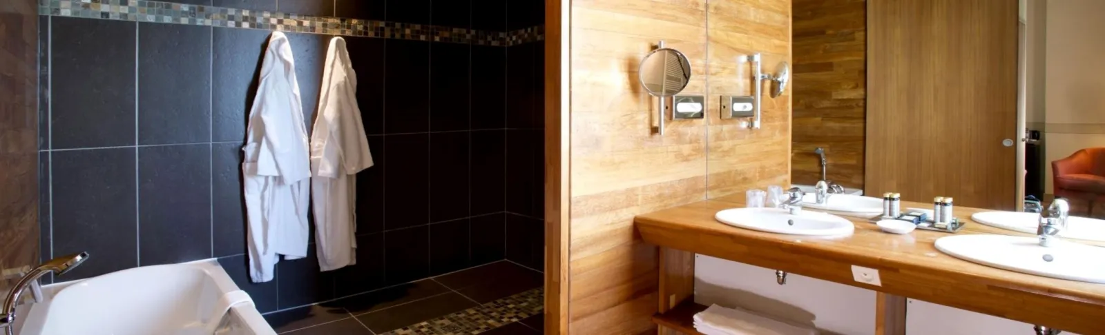 Luxury bathroom at Château Grattequina in Bordeaux featuring dual white marble sinks, round mirror, black tile and natural wood finishes, glass-enclosed shower with white bathrobes