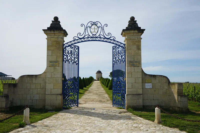 Ornate blue and gold entrance gate of Château Balestard La Tonnelle in Bordeaux, flanked by two stone pillars, leading to a cypress-lined avenue towards the château