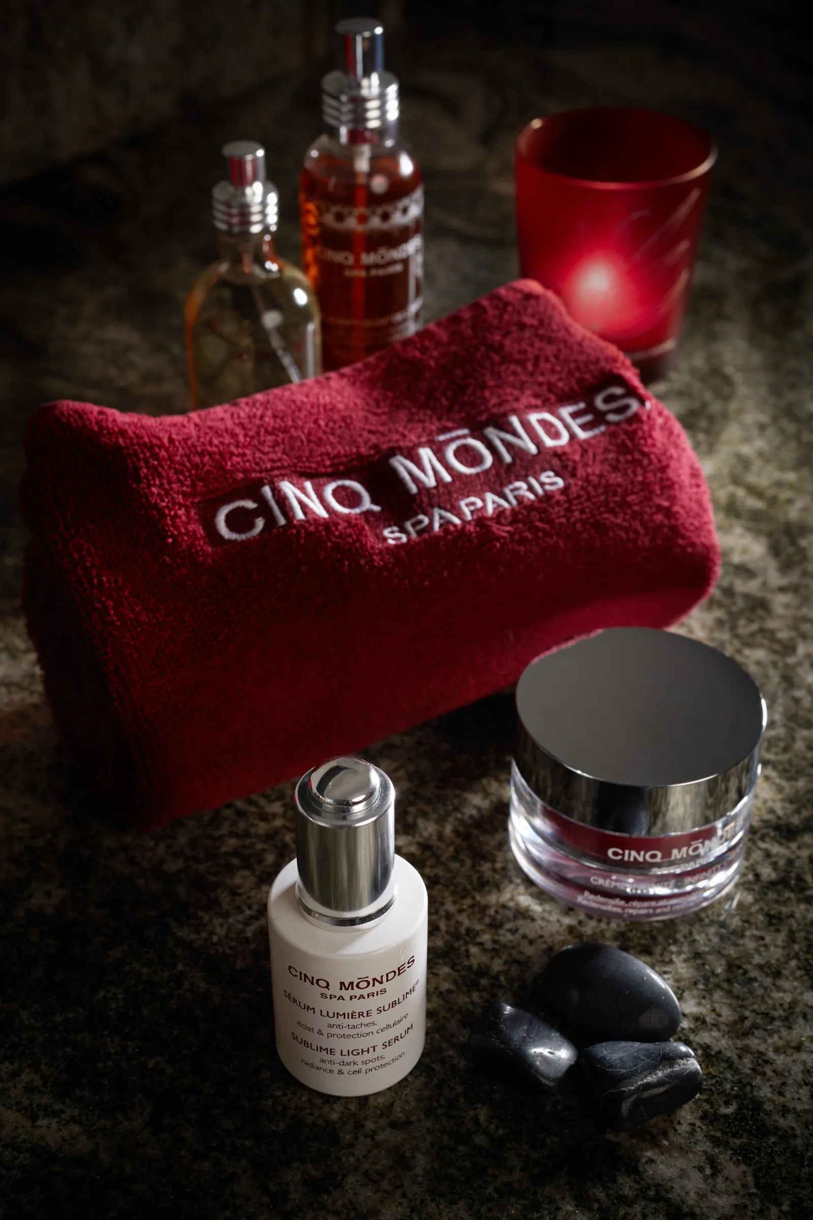 Luxury Cinq Mondes Paris skincare products arranged on grey surface including burgundy towel, fragrance bottles, cosmetic cream jar, black massage stone and glowing red candle for premium spa experience.
