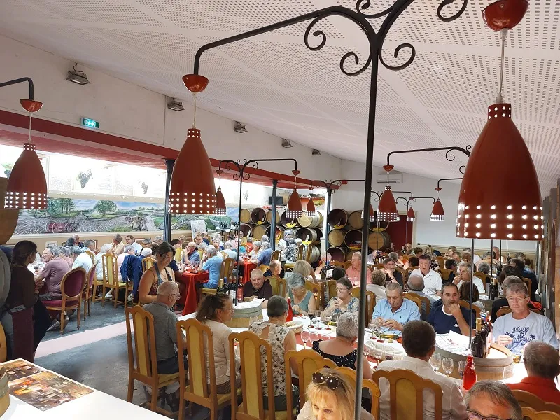 Dining room at Château Marquis de Vauban in Bordeaux filled with guests seated at tables, featuring ornate red iron pendant lights and panoramic vineyard views through large windows.