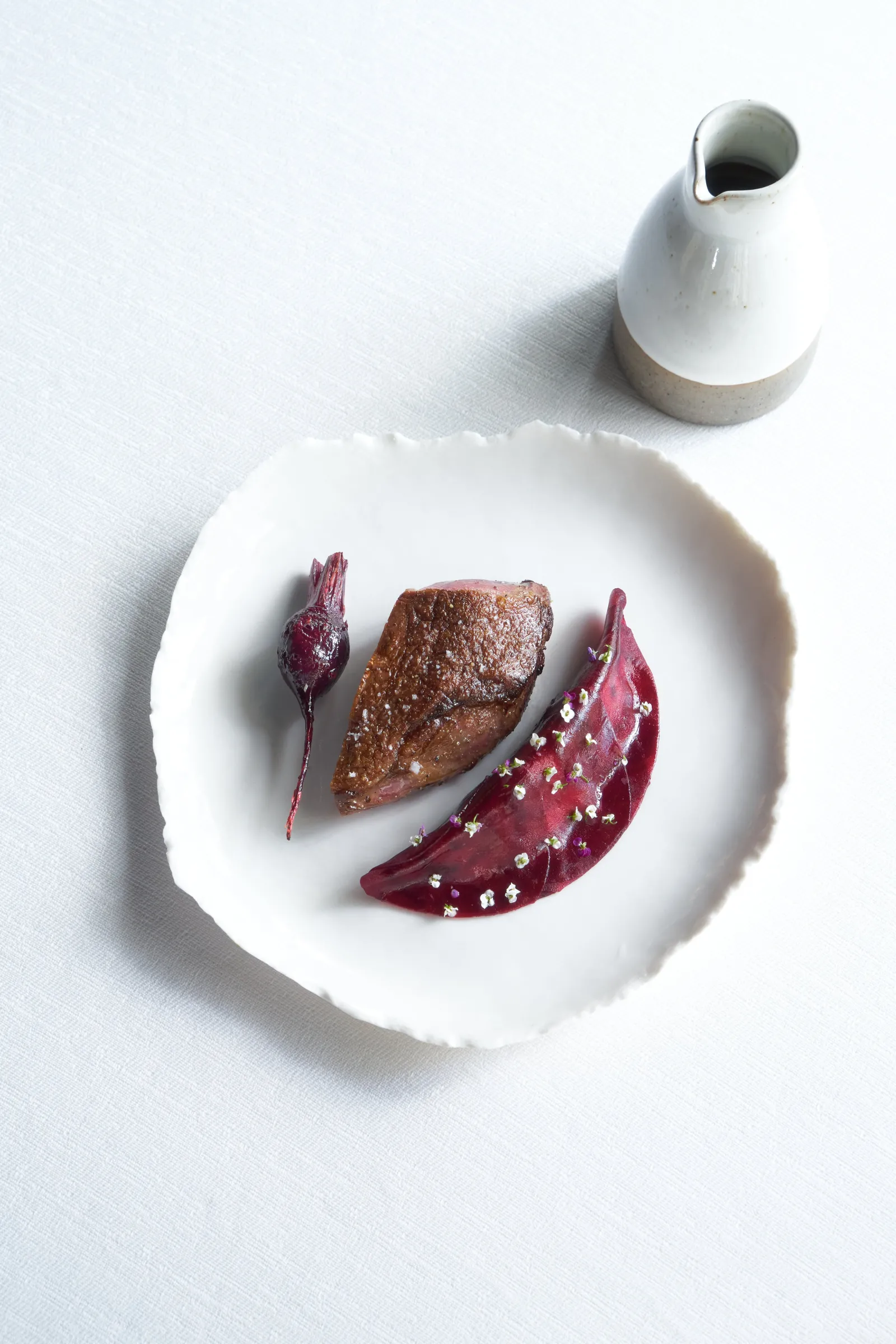 White plate with seared foie gras accompanied by red beetroot slices and garnish, served with a white porcelain pitcher