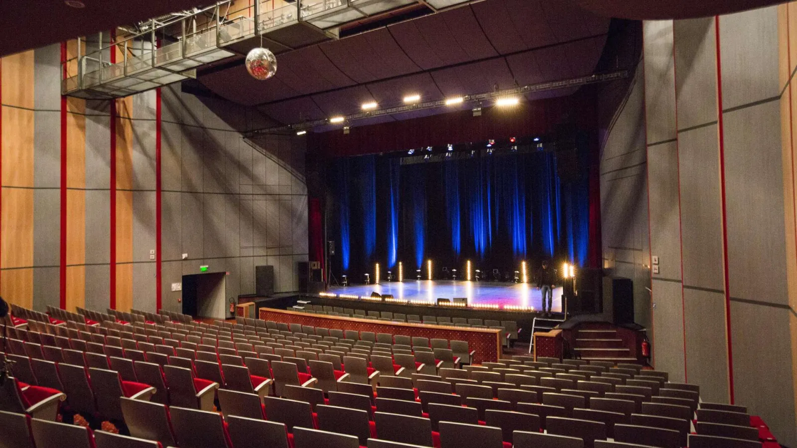 Interior of a modern theater auditorium with red tiered seating, illuminated stage with blue curtains and colored lighting, walls decorated with vertical beige and red panels.