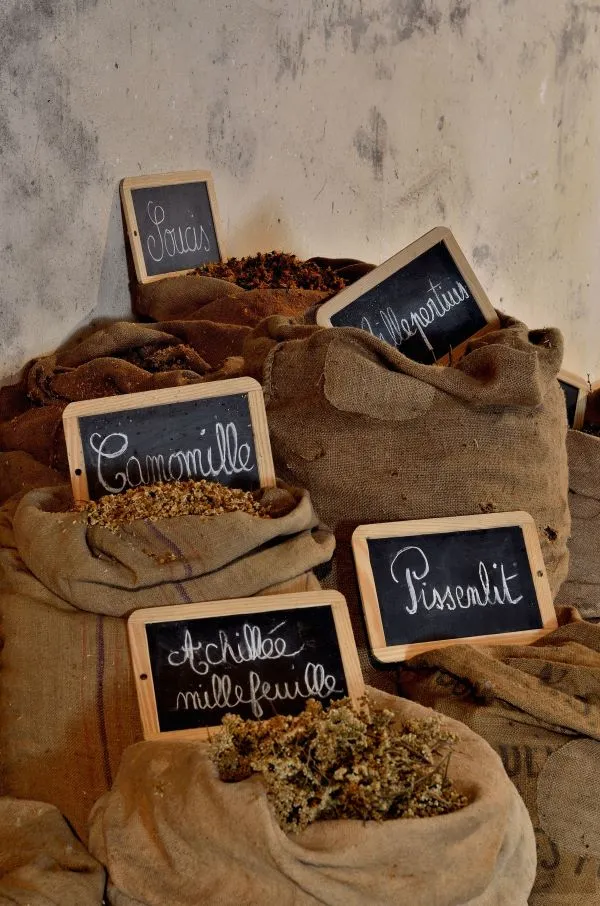Burlap sacks filled with various herbs and spices with slate labels displaying their names: Honey, Flower, Chamomile, Yarrow, Horehound, and Dandelion, arranged against a weathered wall.