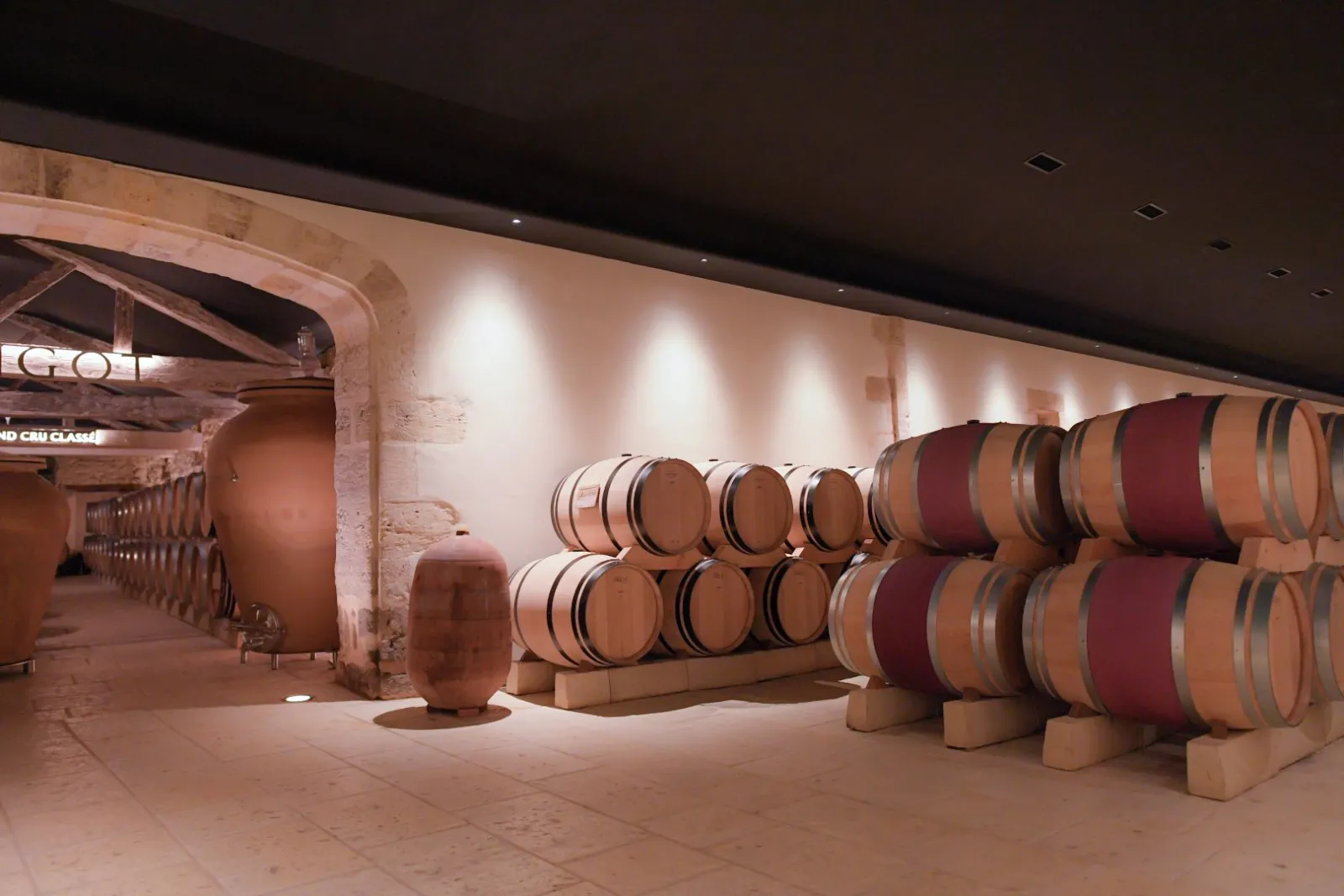 Winery cellar at Château Mangot in Bordeaux featuring rows of oak barrels under modern lighting in a traditional stone building.