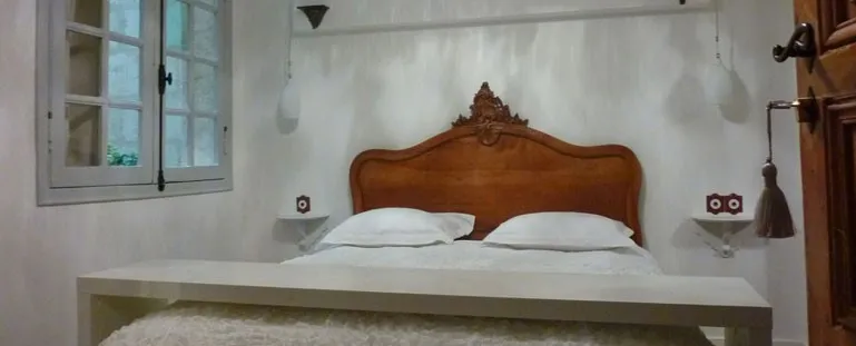Elegant bedroom featuring a carved wooden bed with baroque details, pristine white linens, whitewashed walls, a window with panes, and minimalist décor in classic Bordeaux style.