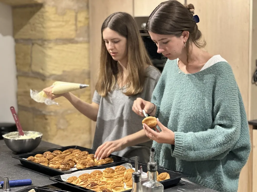 Two women participate in a cooking class in Bordeaux, preparing golden pastries on black baking trays in a professional kitchen.