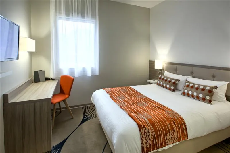 Modern hotel room featuring a double bed with white linens and striped orange throw, desk with orange chair, television, window with white curtains and contemporary lighting