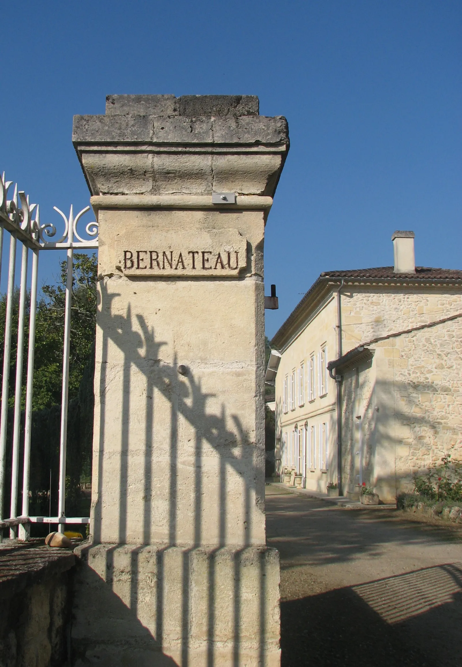 Entrance gate pillar of Château Bernateau in Bordeaux with stone carved name, driveway and traditional Bordeaux building in the background under blue sky