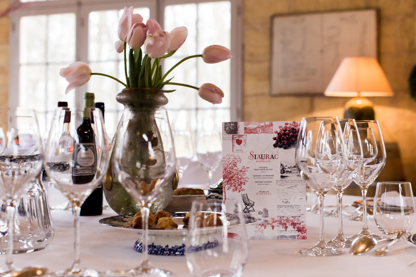 Elegant table setting at La Table de Siaurac restaurant in Bordeaux, featuring wine glasses, pink tulips in a vase, Siaurac menu, wine bottle and table lamp with bright windows in the background