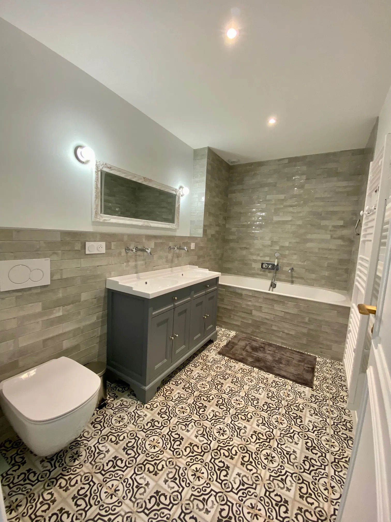 Modern bathroom featuring a black vanity sink, wall-mounted toilet, built-in bathtub, geometric black and white tile flooring, gray stone wall tiling, illuminated mirror, and ceiling spotlights.