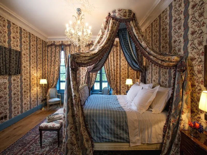 Elegant bedroom at Château Soutard in Bordeaux featuring a canopy bed with ornate draped fabrics, floral patterned wallpaper, crystal chandelier, and classic furnishings.