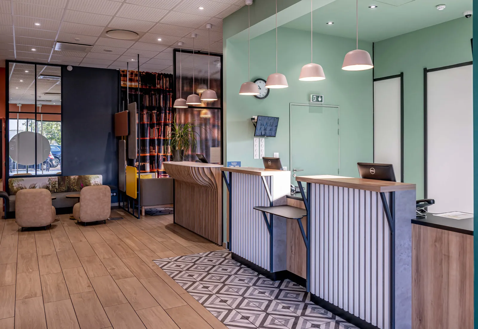 Modern reception area of B&B Hotel Bordeaux Bassins-à-Flot featuring wooden counters, mint green walls, pink pendant lights, seating area with beige armchairs, bookshelf, light wood flooring and black glass partitions.