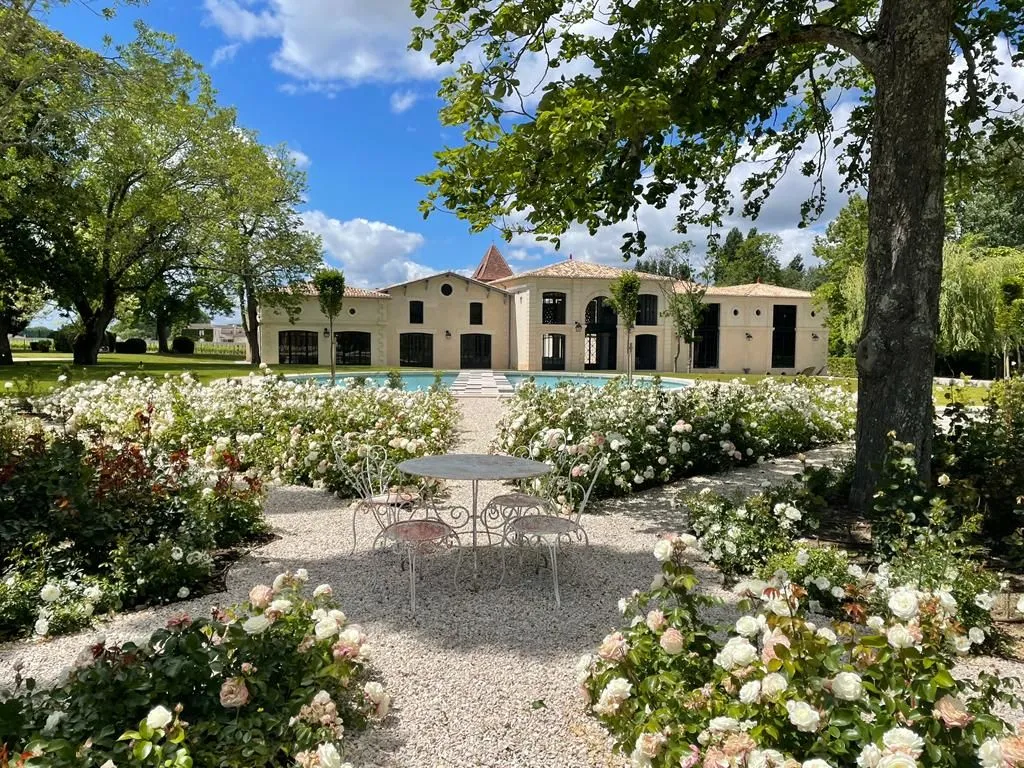 Château Montlabert in Bordeaux: cream-colored stone mansion with tiled roof surrounded by manicured gardens featuring white and pink flowers, ornate metal terrace, and mature trees.