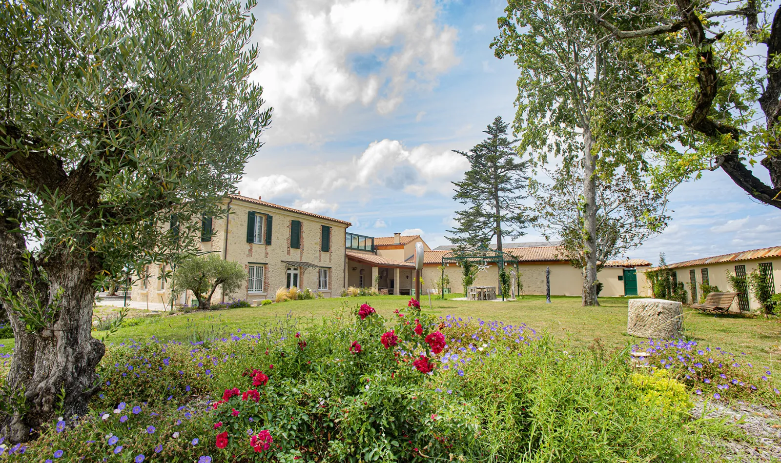 Panoramic view of Domaine des Perchés near Bordeaux featuring stone buildings, a flowering garden with red roses and blue flowers, tree-lined pathways with ancient olive and shade trees against a cloudy sky.