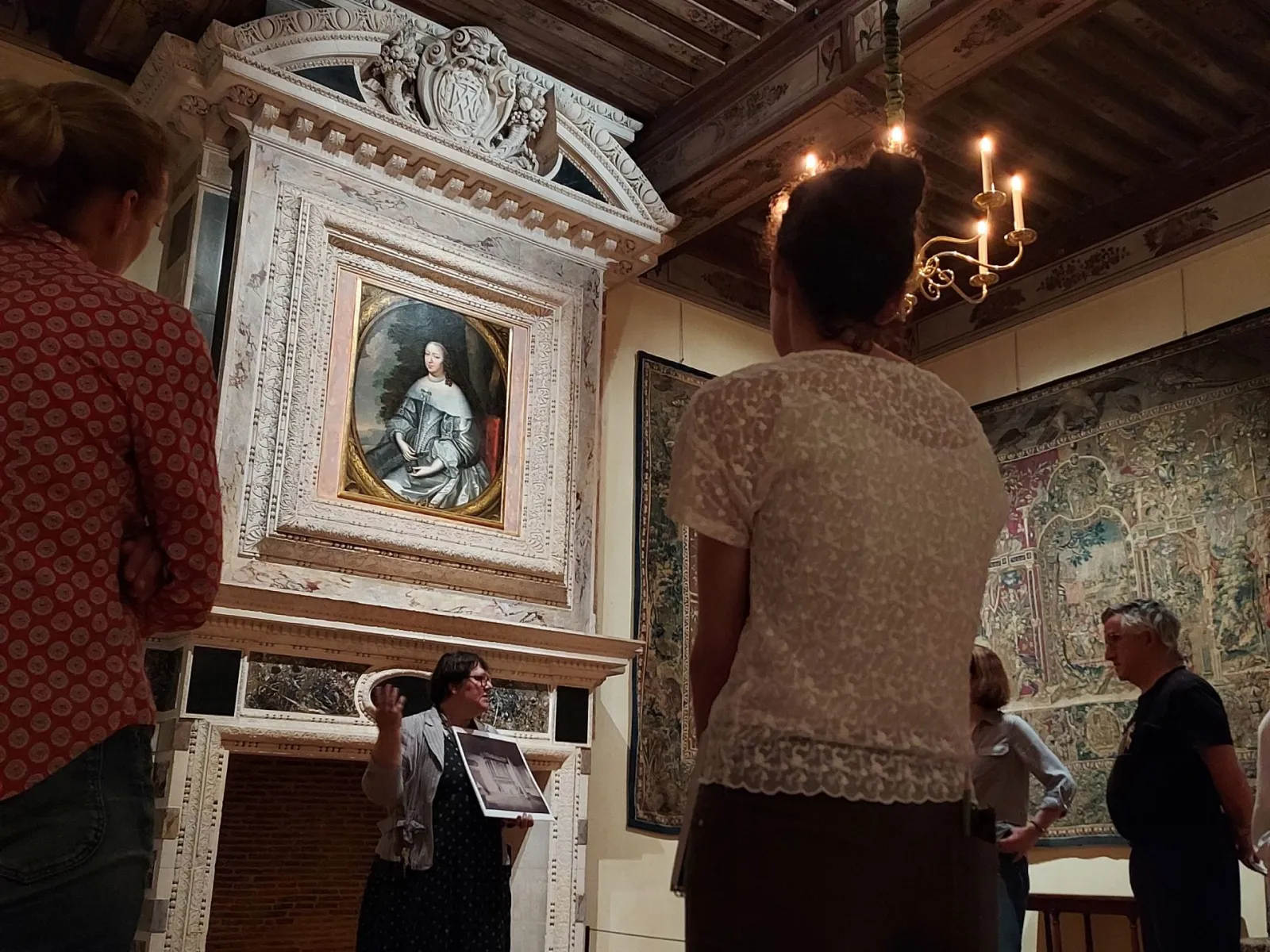 Guided tour in a room of Cadillac Castle with visitors viewing a framed portrait of a 17th-century aristocratic woman, wall tapestries, and a candelabra chandelier.