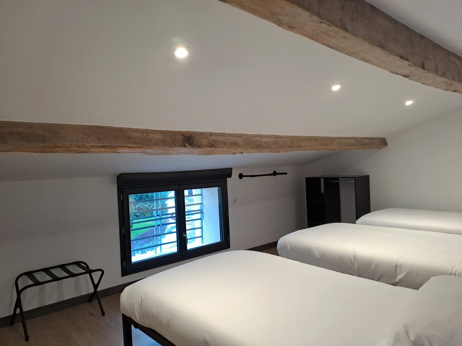 Spacious bedroom in Anthocyane guesthouse featuring two white beds, exposed wooden beams, large panoramic window with black shutters, recessed lighting and minimalist decor