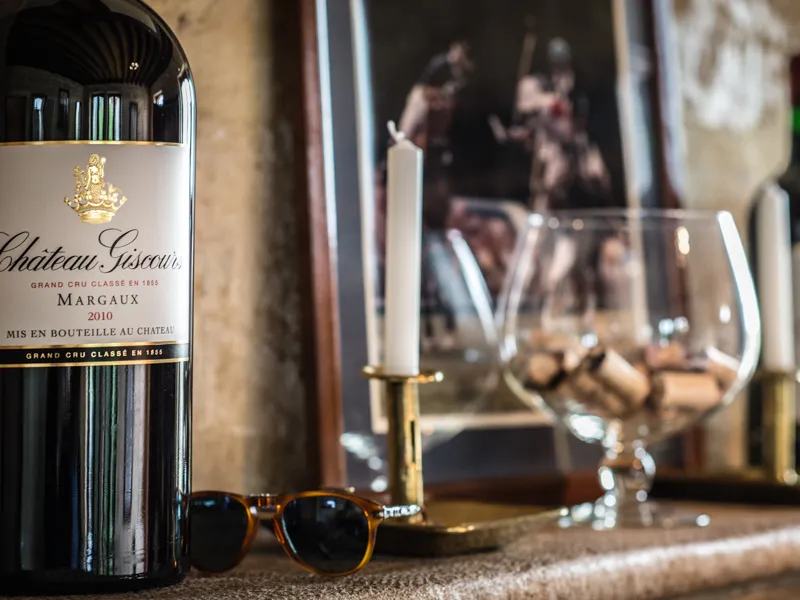 Bottle of Château Giscours 2010 Margaux Grand Cru Classé wine, sunglasses, and empty wine glasses on a table with people blurred in the background of a tasting room.