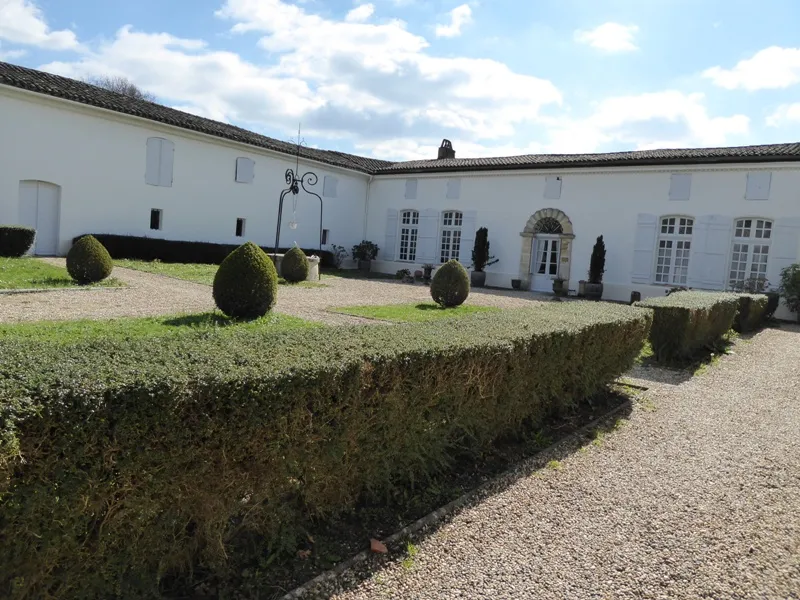 Interior courtyard of Château de Lisennes in Bordeaux featuring white buildings, tiled roofs, manicured boxwood hedges and spheres, gravel pathway and cloudy sky