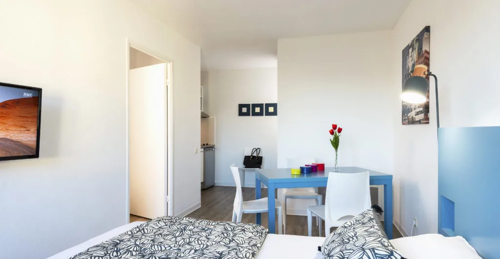 Modern hotel room featuring two twin beds with patterned sheets, blue dining table with white chairs, red flowers in vase, kitchenette in background, and minimalist décor