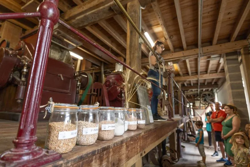 Interior of Moulin de Porchères in Bordeaux featuring glass jars of grains displayed on a wooden table, a red pump in the foreground, and visitors exploring the historic mill machinery in the background.