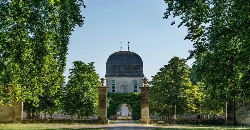 View of Château de Sales in Bordeaux featuring its distinctive blue dome, framed by a tree-lined avenue and bordered by stone porticos.
