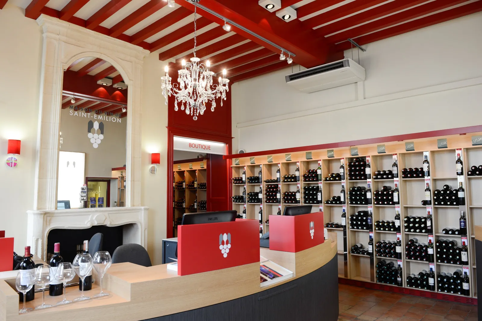 Elegant interior of the Saint-Emilion Wine House in Bordeaux, featuring a wooden and red tasting counter, wine shelves, red and white beamed ceiling, crystal chandelier, and white fireplace.