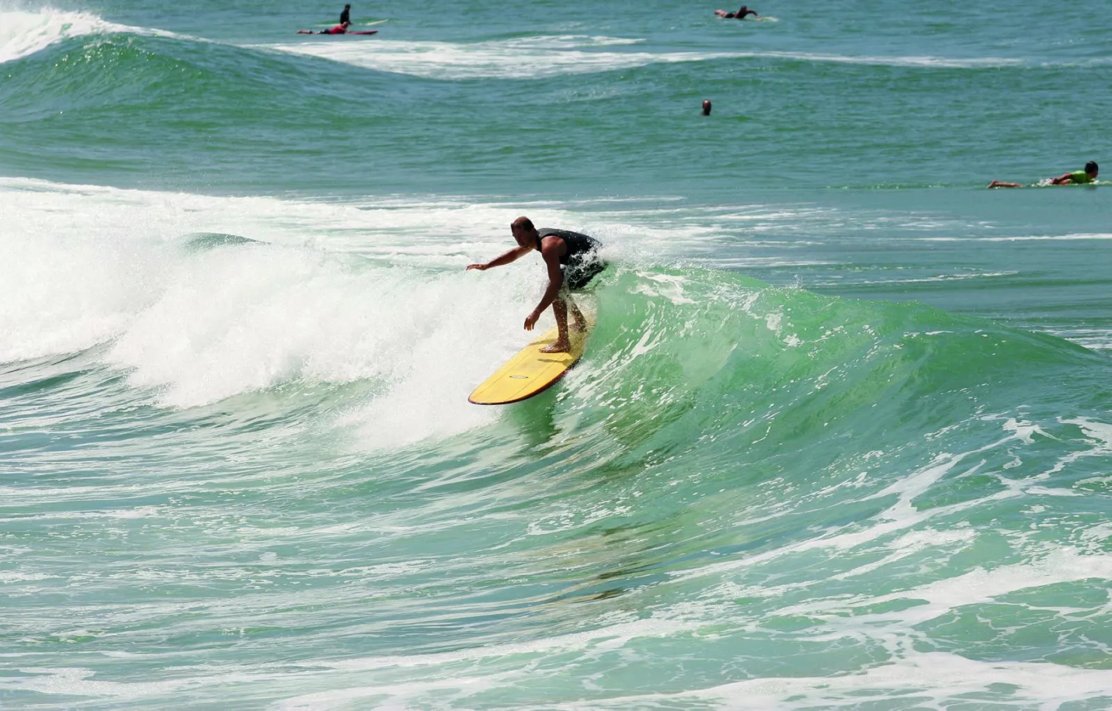A surfer on a yellow board riding a green wave with white foam, surrounded by other surfers in the ocean