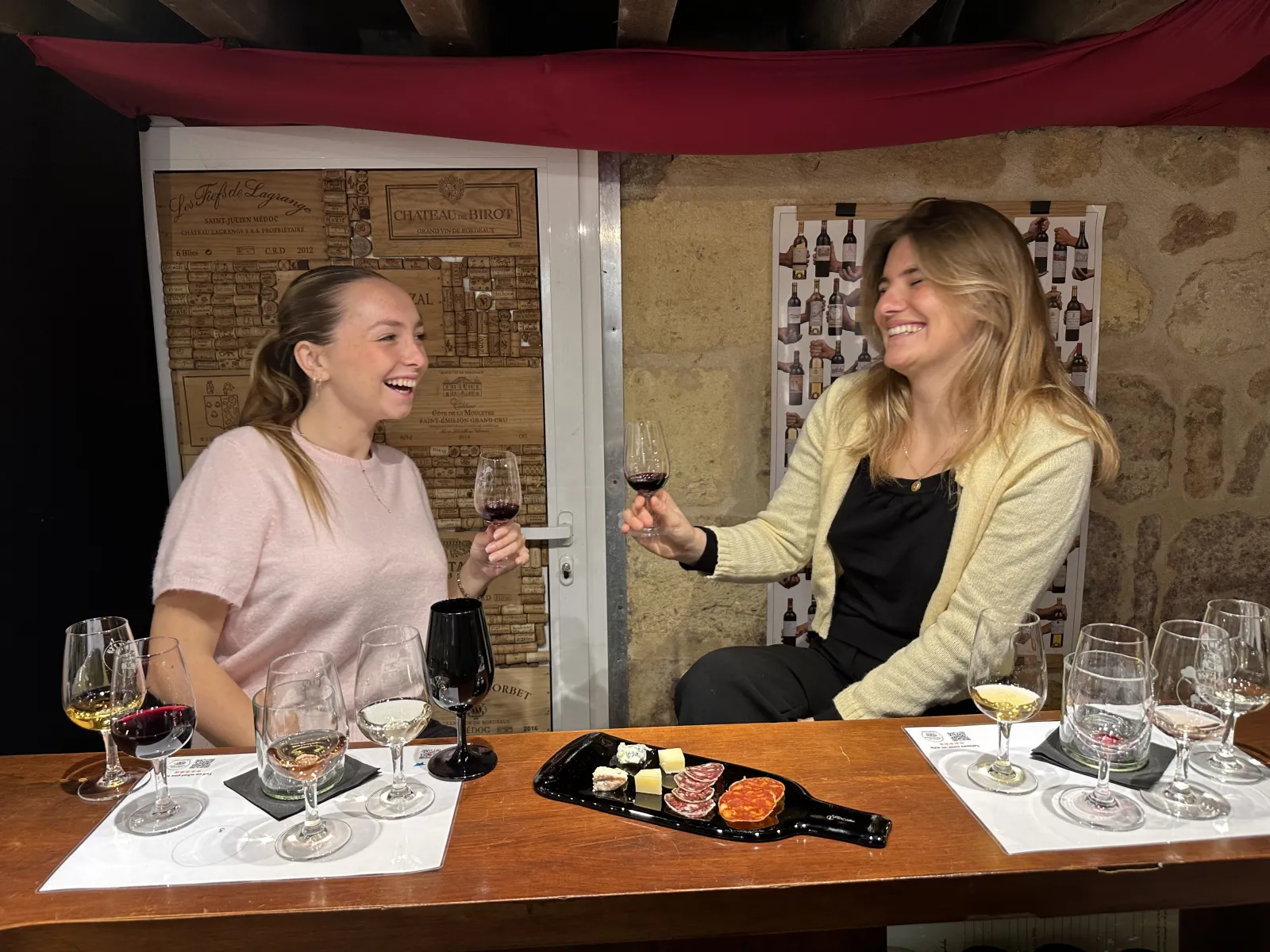 Two smiling women taste red wine in Bordeaux during an I Love my 2CV wine tasting. They sit at a table with wine glasses, tasting devices, and a charcuterie board. Wine posters are visible in the background on stone walls.