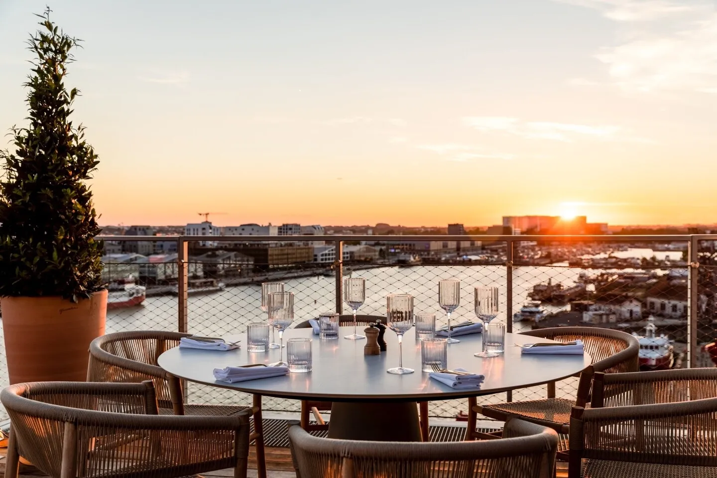 Sunset rooftop terrace with round dining table, rattan chairs, glassware and place settings overlooking Bordeaux cityscape and waterfront