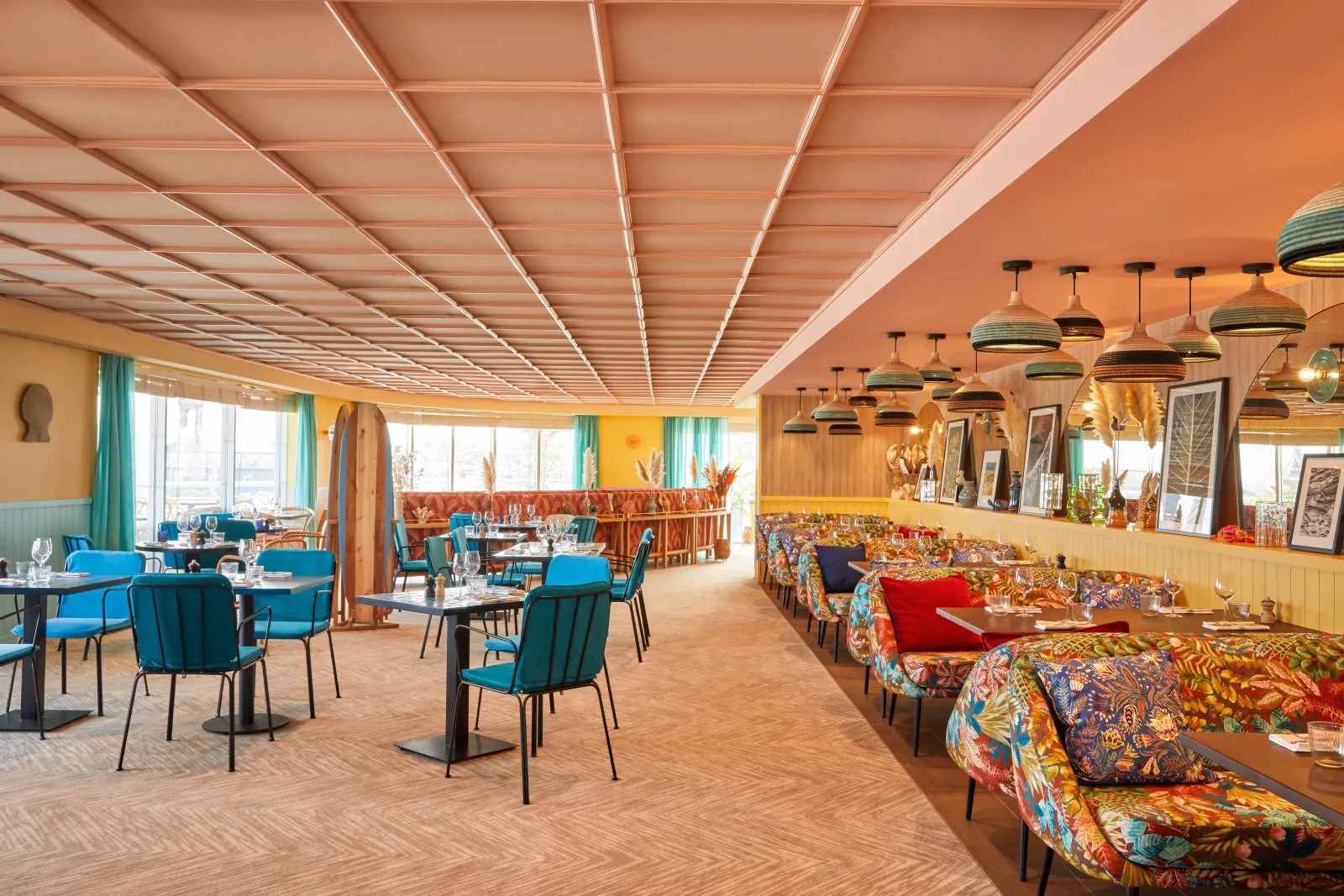 Modern restaurant interior featuring a geometric wooden coffered ceiling, round dining tables with blue chairs, colorful patterned banquettes on the right, black and gold pendant lights, and large windows with warm orange and turquoise color scheme.
