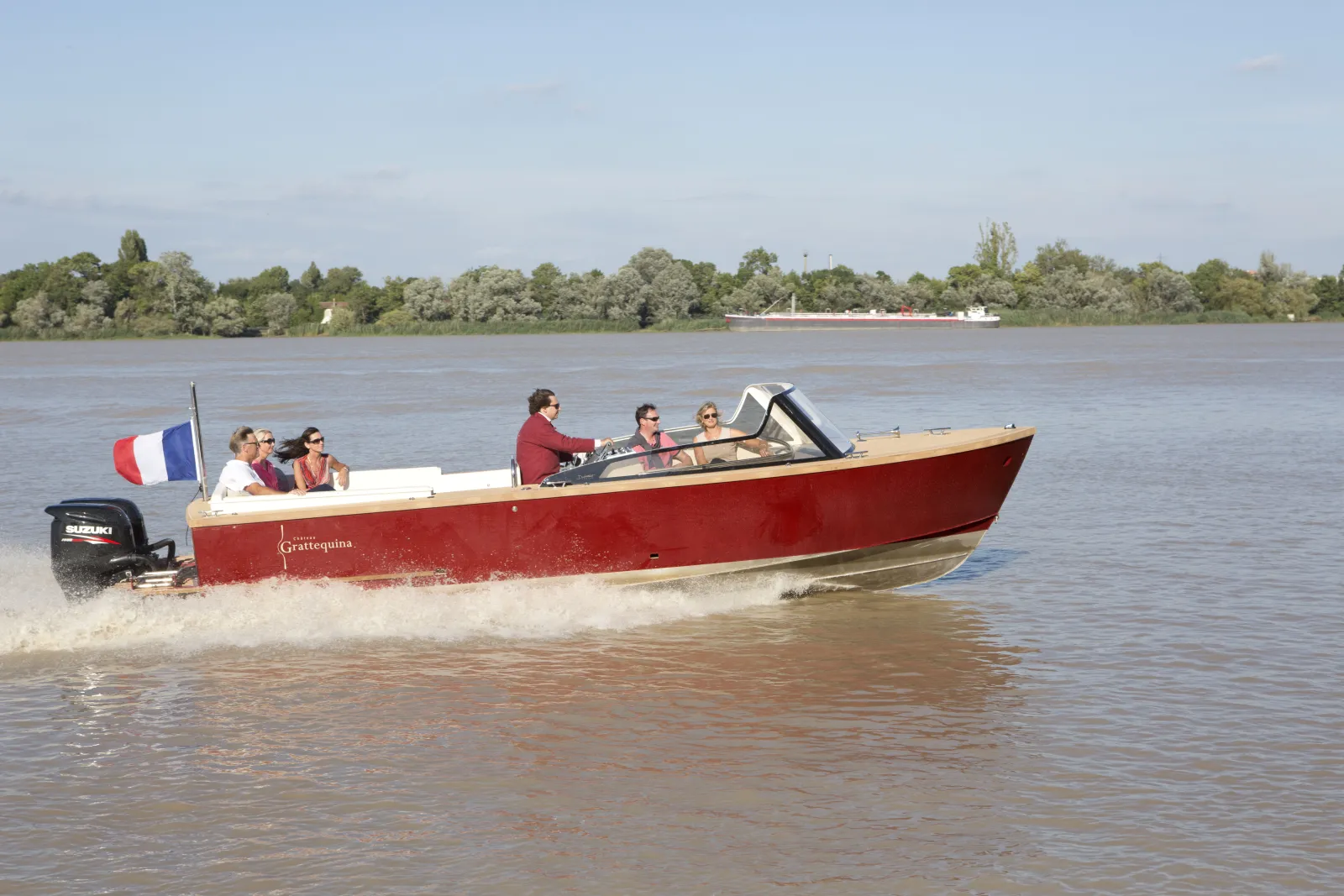 The Grattequina water taxi, a red and white boat, carries passengers on the Garonne River in Bordeaux, with a French flag on board and wooded banks in the background.