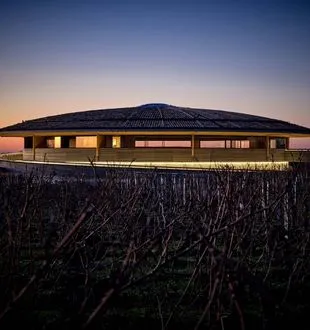 The Dome of Bordeaux glowing at dusk, a modern rounded building in a harvested cornfield with lit windows against a blue and orange twilight sky.