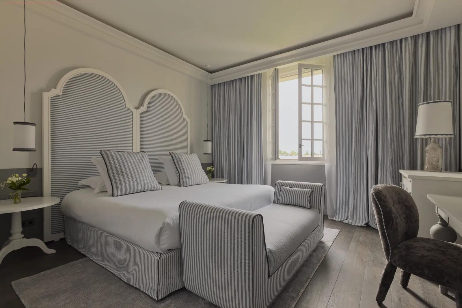 Elegant bedroom featuring a striped grey and white double bed with arched headboard, pearl grey curtains, natural light from window, white furniture and black chair, refined décor in Bordeaux.