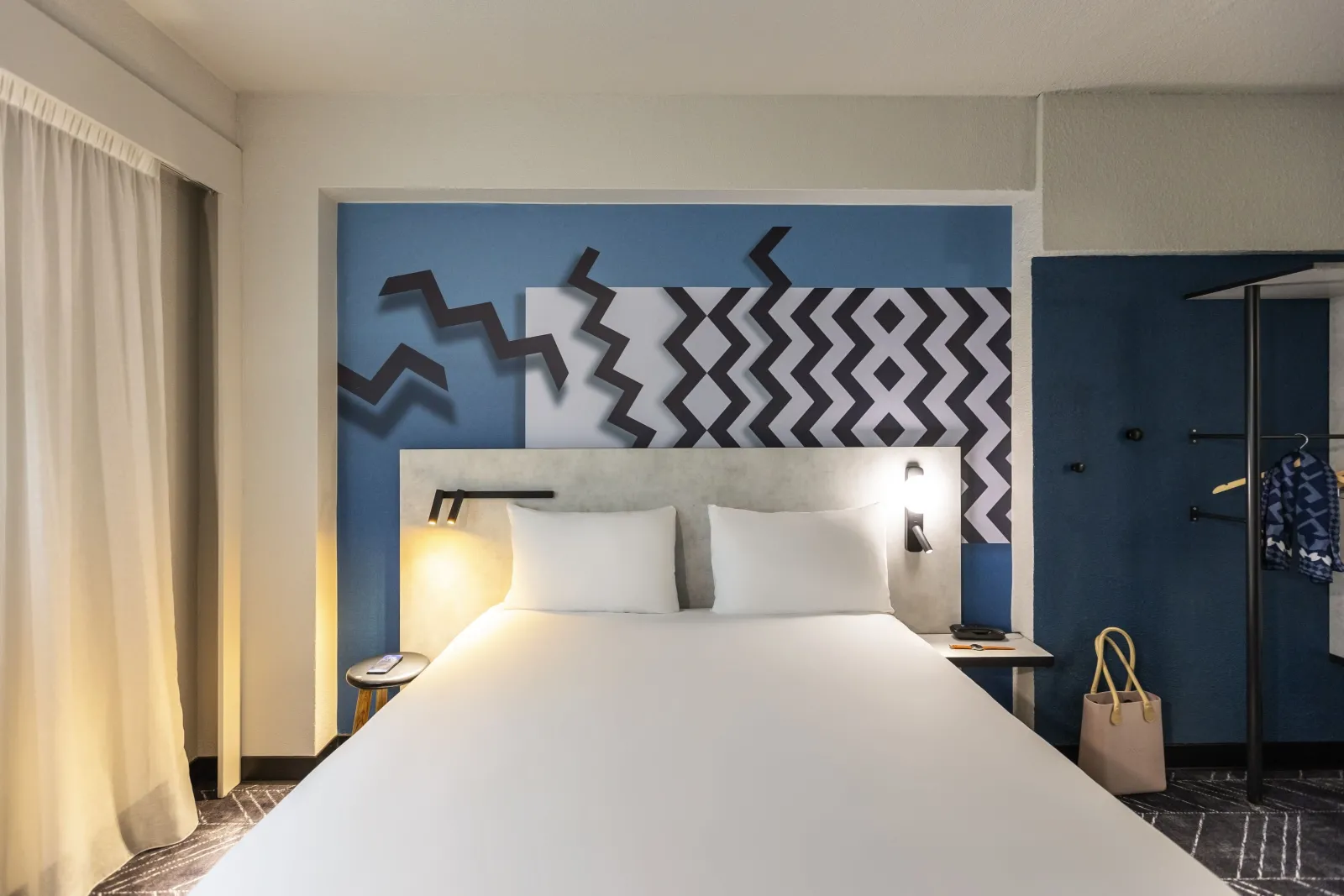 Modern hotel room at Ibis Styles Bordeaux Meriadeck featuring a white bed, blue and grey geometric patterned headboard, ambient lighting, and contemporary furnishings