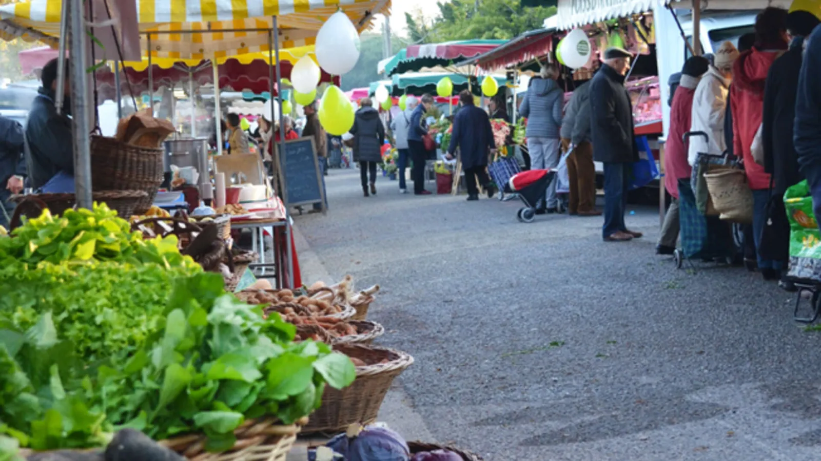 View of Gradignan market in Bordeaux featuring fresh produce stalls under colorful awnings, shoppers browsing between stands, and yellow decorative balloons.
