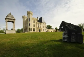 Old Lormont Castle in Bordeaux featuring crenellated stone towers surrounded by green lawns and contemporary sculptures under a partly cloudy sky
