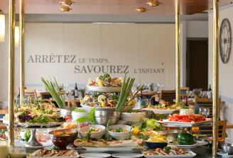 Gourmet buffet display featuring seafood platters, fresh salads, fish, and colorful vegetables in a restaurant overlooking Bordeaux. French motto on wall encouraging to savor the moment.