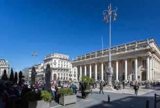 The Grand Theatre of Bordeaux, a neoclassical building with majestic white columns, ornamental façade and clock, overlooking a bustling public square under clear blue sky.