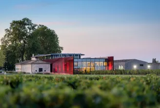 Château La Dominique in Bordeaux: modern architecture with red and grey facades integrated into vineyards, historic white building in background and centuries-old oak tree.