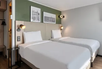 Modern double bedroom at Campanile Nature Bordeaux Sud Pessac hotel featuring two beds, beige headboard, sage green and grey walls, black and white photographs, brass wall lights and contemporary furniture.